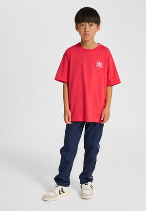 Rød kortærmet t-shirt med et logo på brystet, kombineret med mørkeblå træningsbukser og beige sneakers med sorte striber.