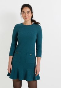 Vestido texturizado en color teal con mangas largas, escote redondo, dos bolsillos frontales con detalles dorados y un dobladillo acampanado con volantes.