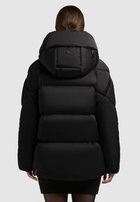 Selja näidatud inimene mustas puhv-jope ja suure kapuutsiga, seljas must seelik, lihtsa heleda halli tausta ees.