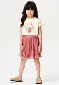 Crème graphic t-shirt met een ontwerp van vogels en een hart, gecombineerd met een geplooide roze rok en beige instap schoenen. Eenvoudige en casual outfit.