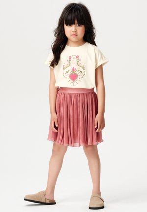 Crème graphic t-shirt met een ontwerp van vogels en een hart, gecombineerd met een geplooide roze rok en beige instap schoenen. Eenvoudige en casual outfit.