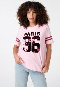 Lichtroze katoenen t-shirt met korte mouwen, met een zwart en roze paillettenafbeelding met de tekst "PARIS 36" en twee bordeauxrode schouderstrepen.