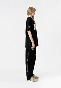 Camiseta negra de gran tamaño con estampado gráfico blanco, combinada con pantalones negros que tienen detalle de cremallera lateral y zapatos marrones con cordones en color azul verdoso.