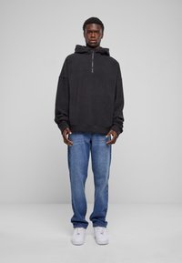 Sort zip-up hættetrøje med en stor hætte, parret med lyseblå jeans og hvide sneakers. Har afslappet pasform og ribbede manchetter.