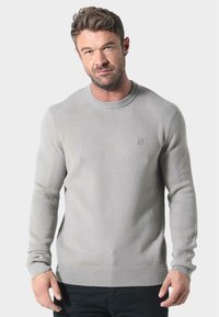 Helles grauer Rundhalspullover aus weichem Stoff, mit ribbeltem Ausschnitt und Bündchen, versehen mit einem kleinen Logo auf der Brust.