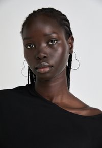 Top nero con scollo asimmetrico; il modello ha i capelli intrecciati e indossa grandi orecchini a cerchio argentati a forma di cuore. Texture della pelle liscia.