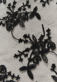 Sort broderet blomsterdesign med detaljerede mønstre på en lys grå stofbaggrund, der viser forskellige blomsterformer og -mønstre.