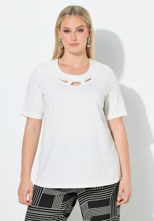 Kvinde med langt blondt hår iført hvid kortærmet top med udskæring ved halsen og sort-hvide bukser med geometrisk mønster.