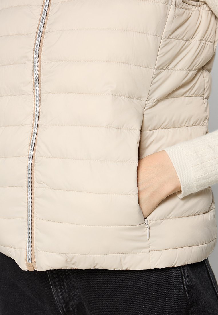 Beige gepolsterte Weste mit horizontalen Nähten, Frontreißverschluss undseitlichen Taschen. Weiche Textur, leichtes Material und minimalistisches Design.