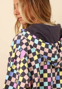 Veste à capuche avec un motif à carreaux multicolores, comprenant des carrés noirs, roses, bleus, jaunes et menthe. Comprend un détail de patch logo rose.