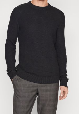 Pull en tricot noir avec un motif texturé, col rond et poignets côtelés. Associé à un pantalon gris à carreaux, présentant une coupe décontractée.