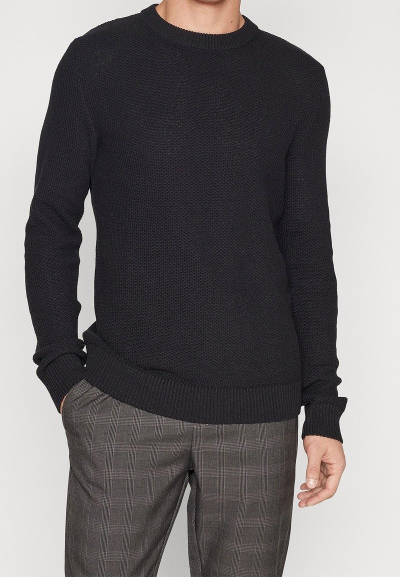 Pull en tricot noir avec un motif texturé, col rond et poignets côtelés. Associé à un pantalon gris à carreaux, présentant une coupe décontractée.