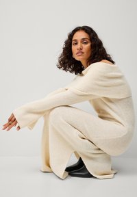 Roomkleurige geribbelde gebreide jurk met een off-the-shoulder ontwerp, lange mouwen, zijsplits en zwarte enkellaarsjes. Glad oppervlak.