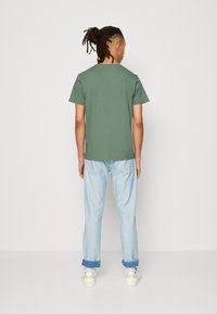 Zielona koszulka z krótkim rękawem z bawełny, w połączeniu z jasnoniebieskimi dżinsami z podwiniętymi mankietami i białymi trampkami, widziana z tyłu.