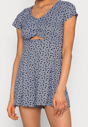 Femme portant une robe bleu marine à manches courtes avec des motifs de marguerites blanches et une petite découpe sur le ventre juste en dessous de la poitrine.