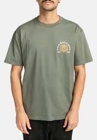 Olijfgroene katoenen T-shirt met korte mouwen. De voorkant heeft een grafisch logo-ontwerp met een leeuw en de tekst "RVCA EST. MMI". Classic fit.