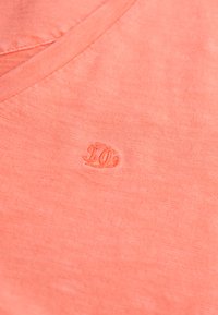 Camiseta coral con cuello en V, confeccionada en suave algodón de textura lisa. Presenta un logo bordado sutil en color a juego en el pecho.