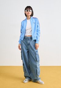 Jeune femme portant un crop top blanc, une veste de survêtement Puma bleu clair, un jean cargo bleu large, des baskets argentées et des lunettes de soleil blanches, debout sur un sol jaune.