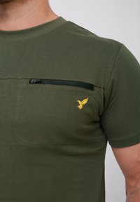 Olivengrøn kortærmet t-shirt med en brystlomme, der har en sort lynlås, og et lille gult fuglelogo broderet på lommen.