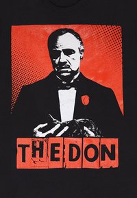 Černé tričko s grafickým designem muže v smokingu na červeném pozadí s textem "THE DON" v oranžových blokových písmenkách.