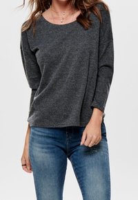 Vrouw in een donkergrijze, langetermijngaren trui met een textuur en een blauwe vervaagde spijkerbroek, met gelaagde kettingen en los haar.