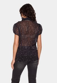 Črna prosojna bluzica z belimi vzorci v obliki zvezd, nabranimi rokavi in nabrano ovratnikom. Oblečena z temno sivo jeans hlačami.