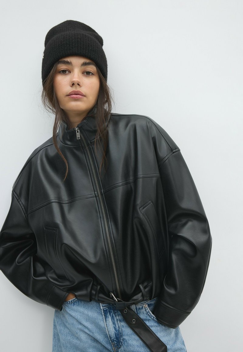 Jeune femme portant un bonnet noir et une veste en cuir noire oversize avec fermeture éclair et ceinture, debout devant un fond clair uni.