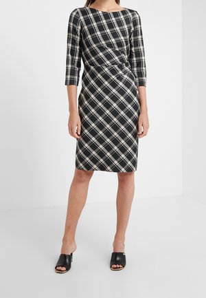 Kobieta ubrana w czarno-białą kraciastą sukienkę do kolan z rękawami do połowy przedramienia oraz czarne, otwarte na palce mules na obcasie, stojąca na białym tle.