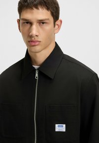 Giacca nera con zip e colletto, due tasche frontali e etichetta del marchio in evidenza. Tessuto dalla trama liscia, vestibilità sartoriale e design moderno.