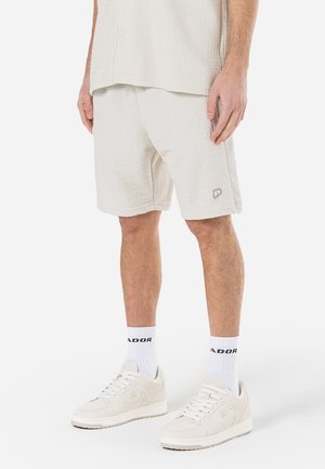 Modèle masculin portant un short crème texturé, une chemise assortie, des chaussettes blanches avec un texte noir, et des baskets blanches décontractées, debout sur un fond blanc.