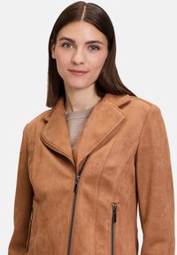 Tan suede jacket with zippered front and pockets, featuring a notched collar and a fitted design. Soft texture with smooth zippers.