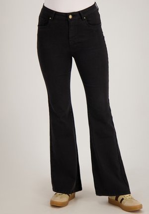 Persoon draagt zwarte high-waist wijde jeans en beige sneakers met bruine strepen, staand tegen een effen achtergrond.