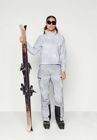 Kvinne i grå skiklær og svarte skistøvler holder et oppreist par rosa og svarte ski mot en enkel hvit bakgrunn.