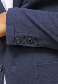 Manschett av en marinblå kavaj med fyra svarta knappar, som visar detaljerad sömnad och en del av en slag ficka.