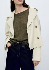 Giacca beige doppiopetto con grandi bottoni, indossata sopra un top verde oliva strutturato e jeans blu scuro, con un classico design a gamba dritta.