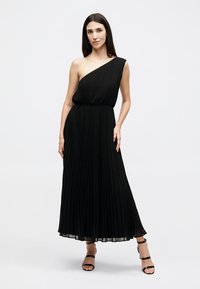 Γυναίκα με μακριά σκούρα μαλλιά, ντυμένη με μαύρο, πλισέ, μάξι φόρεμα με έναν ώμο και μαύρα πέδιλα με λουράκια, στέκεται μπροστά σε απλό λευκό φόντο.