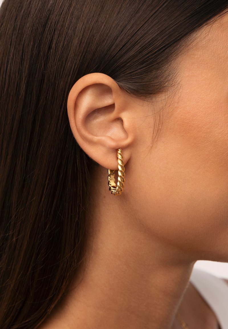 Orecchino ad arco attorcigliato in oro, con un design a spirale testurizzato, indossato all'orecchio di un modello, con capelli e pelle lisci visibili sullo sfondo.
