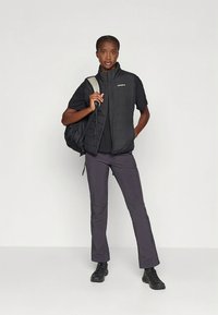 Zwarte geïsoleerde bodywarmer over een zwart shirt, grijze broek en zwarte laarzen. Het model draagt een zwarte rugzak. Eenvoudig ontwerp met een minimalistische uitstraling.