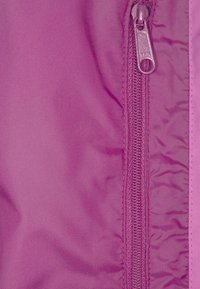 Detalj av en fuchsia nylonjacka med en slät dragkedja med metalldrag och omgiven av ett texturerat tyg med matt yta.