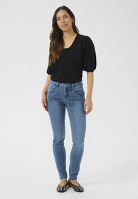 Černý top s balónovými rukávy, modré skinny jeans a černé ploché boty se stříbrnými cvočky. Modelka stojí ve středu před jednoduchým bílým pozadím.