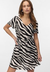 Kvinde iført en kortærmet V-hals kjole med sort-hvide zebrastriber, bundet i taljen, stående foran en ensfarvet baggrund.