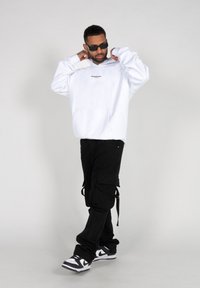 Weißer Kapuzenpullover mit einer Fronttasche und schwarzem Textlogo, kombiniert mit schwarzen Cargo-Hosen mit mehreren Taschen und Riemen sowie schwarz-weißen Sneakers.