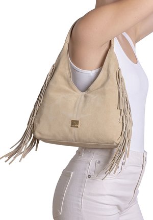 Vrouw met een wit hemdje en broek, draagt een beige suède schoudertas met franjes en een gouden logoplaatje.