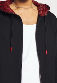 Sudadera negra con cremallera y forro interior rojo con patrón. Presenta cordones con puntas rojas y una camiseta sin mangas blanca debajo. Collares de cadena plateada visibles.