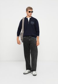Marineblauwe trui met een rits en logo, grijze jeans en een zonnebril. Afgemaakt met witte sneakers met zwarte strepen en een grijze tas.