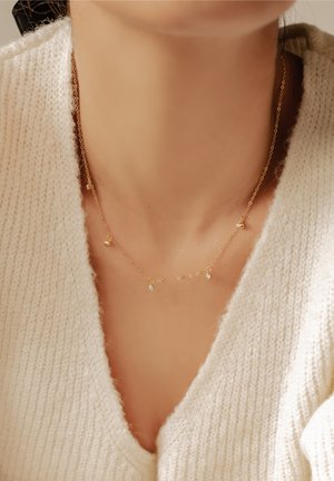 Gold chain necklace with small circular pendants, featuring clear accents. Worn over a soft, cream-colored knitted sweater.