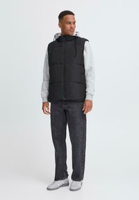 Schwarze gepolsterte Weste mit hohem Kragen über einem grauen Kapuzenpullover. Die Weste hat einen Reißverschluss vorne und eine verstellbare Kapuze, kombiniert mit grauen Jeans und weißen Turnschuhen.