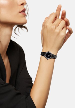 Montre noire avec un cadran rond, un bracelet текстuré et des marqueurs d'heure contrastants. L'aiguille est en position, mettant en valeur le design épuré de la montre.