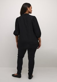 Blusa negra con mangas abullonadas y cuello fruncido. Combinada con unos jeans negros ajustados y zapatos planos. Tejido suave, diseño minimalista.
