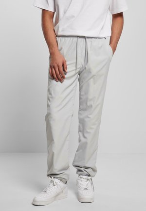 Mann trägt hellgraue Hose mit Kordelzug und Taschen, weiße Sneaker und ein weißes Kurzarmshirt, steht vor einfachem Hintergrund.